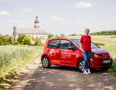 Frau im roten Oberteil vor rotem Auto in der Natur, im Hintergrund eine Kirche