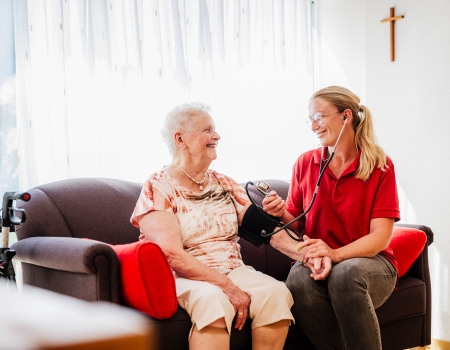 Frau misst Seniorin den Blutdruck, sie sitzen auf einer Couch 